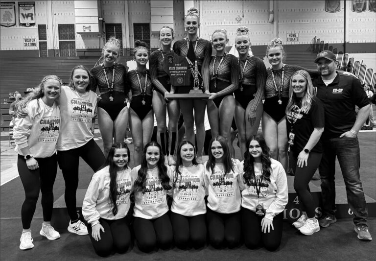 Members of the Dodgeville Co-op Gymnastics Team stand on the podium with its WIAA Division 2 State Championship trophy, the first in the program's history. Team members are (I-r) Coach Kaia Wedig, Coach Elsa Nelson, Abigail Droessler, Myra Barr, Keira Haser, Faith Hoffman, Alaina Nelson, Madelyn Klein, Ezmae Sandberg, Coach Julia Oellerich, and Coach Todd Oellerich; kneeling Left to Right: Elsie Van Overmeer, Natalia Aguirre, Allison Tiber, Bella Smith, and Issie Thompson.