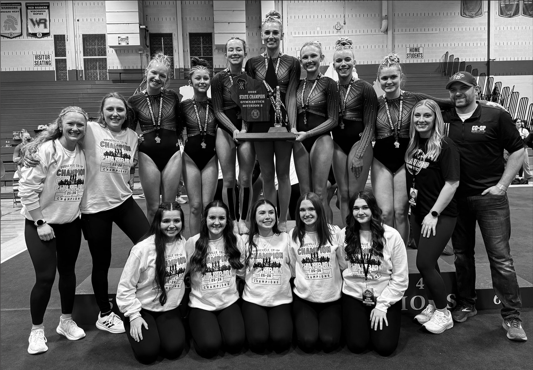 Members of the Dodgeville Co-op Gymnastics Team stand on the podium with its WIAA Division 2 State Championship trophy, the first in the program's history. Team members are (I-r) Coach Kaia Wedig, Coach Elsa Nelson, Abigail Droessler, Myra Barr, Keira Haser, Faith Hoffman, Alaina Nelson, Madelyn Klein, Ezmae Sandberg, Coach Julia Oellerich, and Coach Todd Oellerich; kneeling Left to Right: Elsie Van Overmeer, Natalia Aguirre, Allison Tiber, Bella Smith, and Issie Thompson.